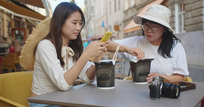 Portrait of happy Asian young female sitting on restaurant terrace and texting on smartphone. Beautiful woman in hat eating chinese food outdoors and talking on street. Leisure concept