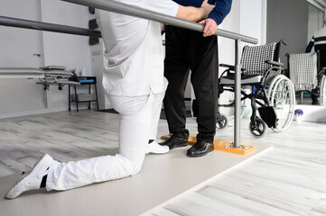 Physiotherapist helping disabled Caucasian senior man to walk with parallel bars in rehabilitation...