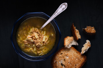 Chicken pasta soup in a blue bowl on a black plate with toasted bread. View from above.