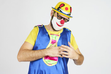 A clown in a bright blue and yellow suit and mask holds his heart with his hands, got sick