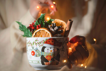 Obraz na płótnie Canvas A young woman holds large mug with full of Christmas flower, oranges, cinnamon sticks and pine cone with lights , close up, still life photography
