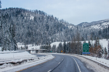 Obraz premium Snow-covered highway among the mountains, on the sides there are trees and trees in the snow. Winter landscape with snow-covered road, fir trees and mountains in Montana, America, 1-18-2020