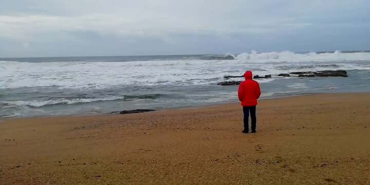 Person wearing a red winter jacket in a beach staring at the ocean