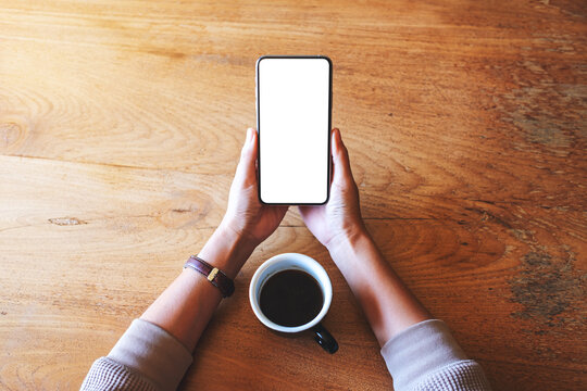 Top view mockup image of a woman holding mobile phone with blank white desktop screen with coffee cup on wooden table