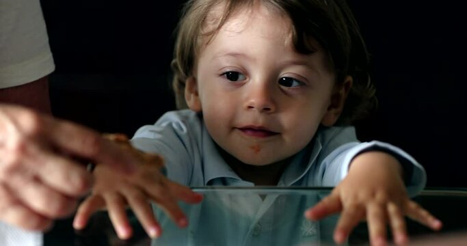 Cute toddler baby boy leaning on glass table with hands peeking, looks at camera