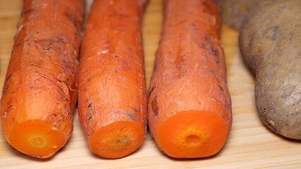 Three boiled carrots close up next to potato with peel prepared to chop on a wooden board