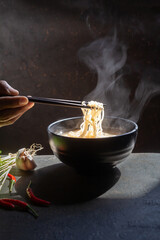 Hand uses chopsticks to pickup tasty noodles with steam and smoke in bowl on wooden background, selective focus. Asian meal on a table, junk food concept