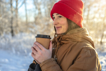 Young woman is drinking hot coffee on a frosty winter Sunny day outdoors in the forest