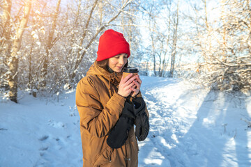 Fototapeta na wymiar Young woman is drinking hot coffee on a frosty winter Sunny day outdoors in the forest