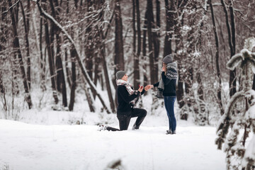 Kneeling guy puts an engagement ring on a girl's hand, making a proposal to get married in winter in a snowy forest