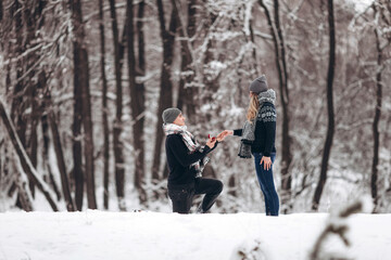 Fototapeta na wymiar A guy kneeling down puts a wedding ring on a girl's hand, making a proposal to marry in a snowy forest in winter