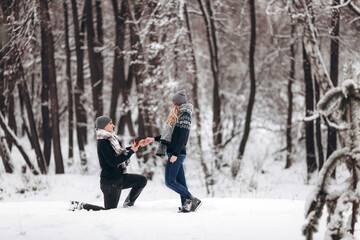 Fototapeta na wymiar A guy kneeling down puts a wedding ring on a girl's hand, making a proposal to marry in a snowy forest in winter