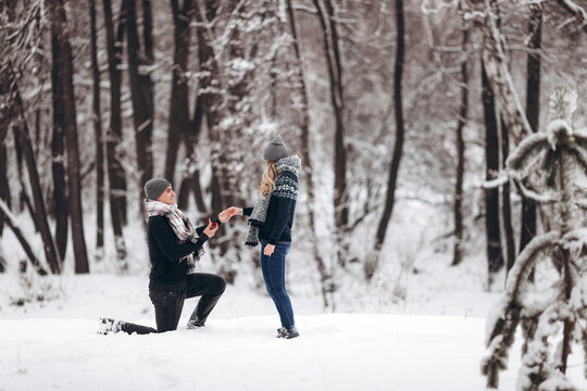 Kneeling guy puts an engagement ring on a girl's hand, making a proposal to get married in winter in a snowy forest