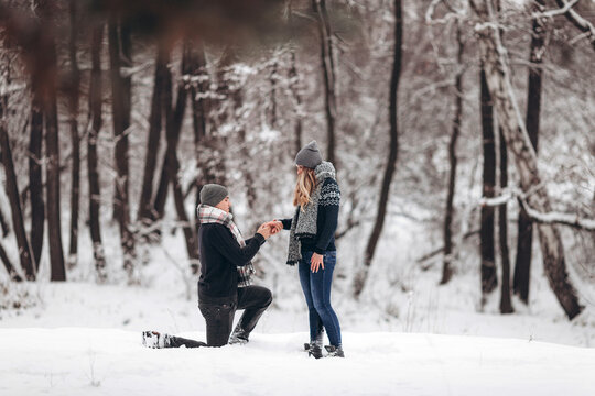 Kneeling guy puts an engagement ring on a girl's hand, making a proposal to get married in winter in a snowy forest