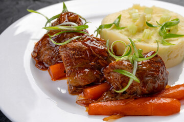 stewed beef pieces in sauce with mashed potatoes and carrots on a white plate. The hot dish is beef...