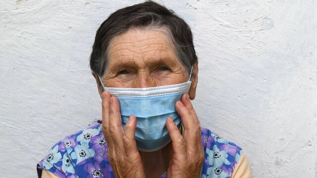 Elderly Woman Touches Single-use Mask With Both Hands. Portrait Of Old Lady With Deep Wrinkles On Her Forehead And Near Eyes. Aged Wrinkled Face Of 80 Year Old Woman