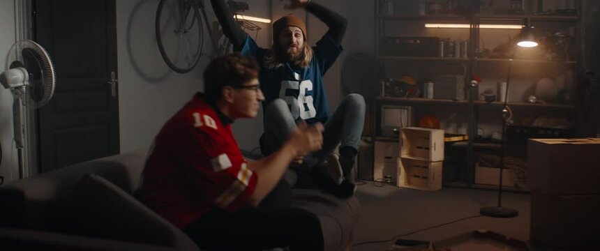 Portrait of two friends playing video game inside garage hideout, winning and celebrating. TV screen is not seen. Shot with 2x anamorphic lens