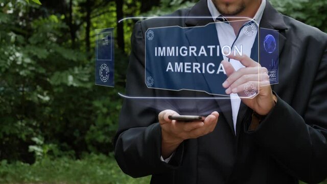 Unrecognizable businessman activates conceptual HUD holograms on smartphone with text Immigration America. Bearded man in white shirt and acket with holographic screen on a background of green trees
