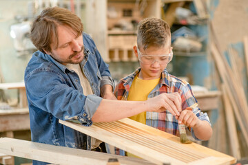 Father teaches his young son how to paint a wooden board with a paintbrush in a carpentry workshop