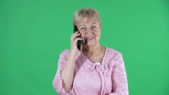 Portrait elderly woman looking at the camera and talking on her smartphone. Gray haired grandmother with short hair in a pink sweater on green screen at studio. Close up.