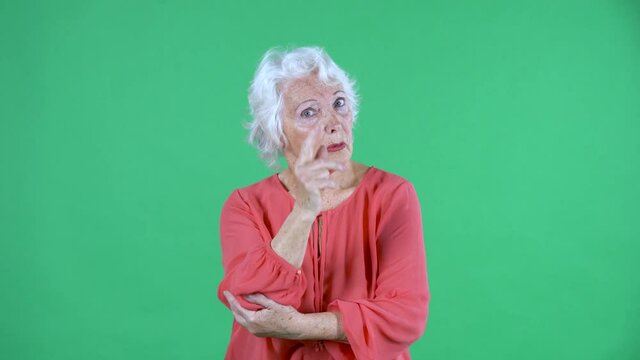 Portrait elderly woman looking at the camera scolding someone and threatening shaking her index finger. Dont do that gesture. Gray haired grandmother in red blouse on green screen at studio. Close up.