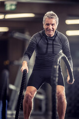 Older man with headphones exercises with ropes in a fitness training center