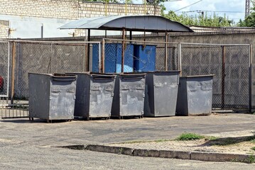 a row of gray iron dumpsters stand on the asphalt outside by the wall of the fence