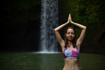 Hands raising up in namaste mudra. Young woman meditating, practicing yoga and pranayama with namaste mudra near waterfall. Yoga outdoor concept. Tibumana waterfall, Bali. Copy space.