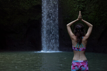 Hands raising up in namaste mudra. Woman meditating, practicing yoga and pranayama with namaste mudra near waterfall. Yoga outdoor concept. Tibumana waterfall, Bali. Copy space. View from back.