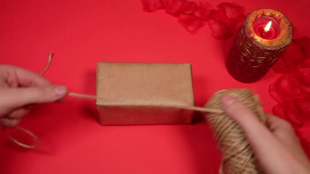 Eco-friendly gift packaging for February 14th valentine's day. A natural rope string. Gift on a red with rose petals, candle. A woman cuts off a rope, sets fire to candles.