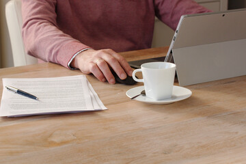 person working at home on a laptop with a cup of coffee, pen and stack of paper on a wooden table background