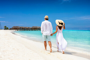Happy couple in white clothing and with hats walks down a tropical beach with turquoise sea in the Maldives islands - obrazy, fototapety, plakaty