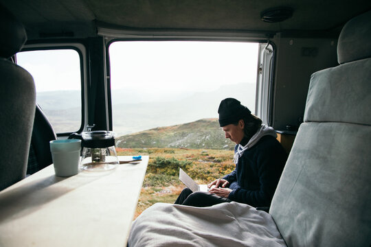 Young man sit inside customised camper van, work on laptop remotely from office, enjoys views while also stays connected. Millennial travel lifestyle. Work remote on the road