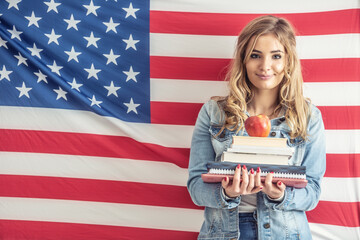 Young teen student holds a stack of study papers and apple on the top with a flag of the Unites States behind her