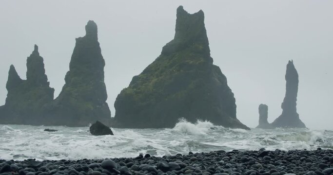 Sharp cliffs on black sand beach of Reynisfjara with waves hitting the shore on foggy rainy stormy day. Vik, Iceland.