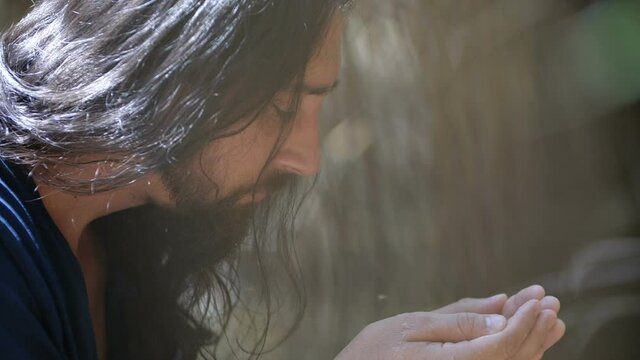 Close up profile portrait thirsty wanderer greedily drinks life-giving water from a stream or lake. A long-haired man in dirty dark clothes  drinks from a handful of water. 200 fps slow motion.