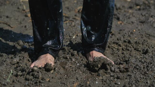 Male bare feet in black pants stand on wet ground, raindrops dripping on top of them. He wiggles his toes funny and his feet are in the mud. 200 fps slow motion