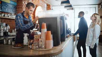 Handsome Male Barista in Checkered Shirt is Making a Latte for a Customer in a Coffee Shop Bar. Beautiful Caucasian Male Cashier Works at a Cozy Loft-Style Cafe Counter in the Background.