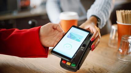 Close Up of a Feminine Hand Holding a Smartphone with an NFC Payment Technology Used for Paying for Take Away Coffee in a Cafe. Customer Uses Mobile to Pay for Latte Through a Credit Card Terminal.