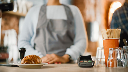Close Up of a Payment Terminal Ready to Pay and Croissant. In Background Beautiful Latin American Female Barista in Coffee Shop Bar. Happy Employee Behind Cozy Loft-Style Cafe Counter in Restaurant.