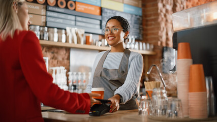 Joyful Multiethnic Diverse Woman Gives a Payment Terminal to Customer Using NFC Technology on Smartphone. Customer Uses Mobile to Pay for Take Away Latte and Pastry to a Barista in Coffee Shop.