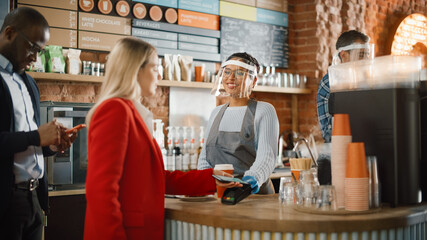 Female Customer Pays for Coffee and Pastry with Contactless NFC Payment Technology on Smartphone to a Barista in Face Shield in Cafe. Social Restrictions Concept in a Bar During Coronavirus Pandemic.