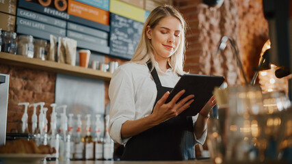 Beautiful Young Caucasian Coffee Shop Manager with Blond Hair is Making Notes on a Tablet Computer and Orders Inventory Items for the Menu in a Cozy Loft-Style Cafe. Successful Restaurant Owner.