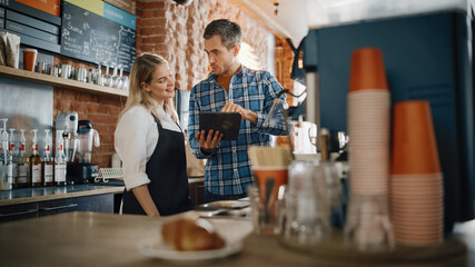 Fototapeta premium Two Diverse Entrepreneurs Have a Team Meeting in Their Stylish Coffee Shop. Barista and Cafe Owner Discuss Work Schedule and Menu on Tablet Computer. Young Female and Male Restaurant Employees.