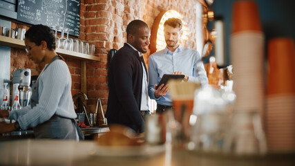 Multicultural Coffee Shop Owners Meeting Behind the Counter and Working on Tablet Computer and Checking Inventory in a Cozy Loft-Style Cafe. Successful Restaurant Managers and Barista at Work.