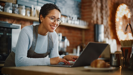 Young and Beautiful Latina Coffee Shop Owner is Working on Laptop Computer and Checking Inventory in a Cozy Cafe. Successful Restaurant Manager Browsing Internet and Accepting Online Take Away Orders.