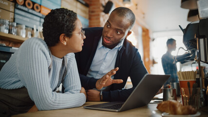 Two Diverse Entrepreneurs Have a Team Meeting in Their Stylish Coffee Shop. Barista and Cafe Owner Discuss Work Schedule and Menu on Laptop Computer. Multiethnic Female and Male Restaurant Employees.