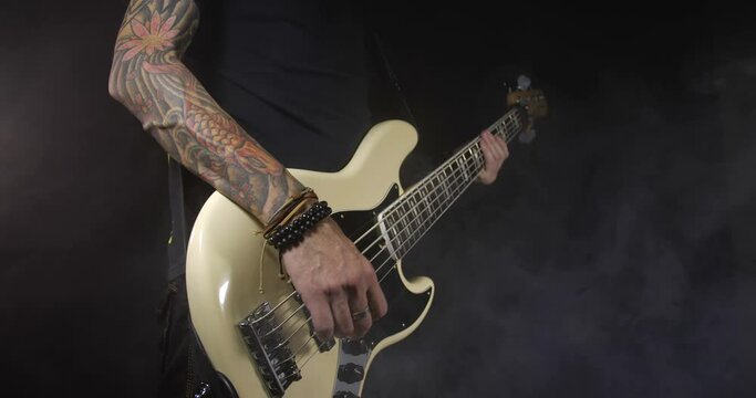 Unrecognizable rock musician with tattoos in black clothes plays ivory bass guitar on black background isolated and smoke