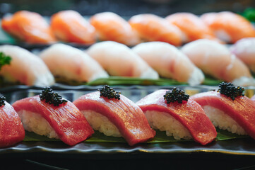 Backlit. Tuna sushi with black tobiko on a black dish. Blurry salmon and hamachi sushi in the background.