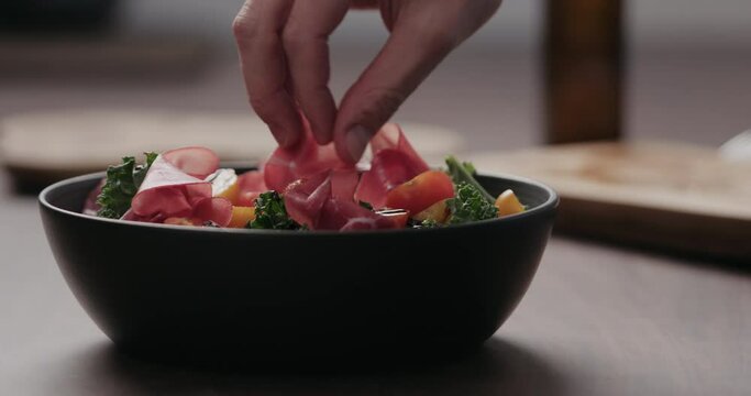 Slow motion man hand add coppa slices to make salad with kale, and cherry tomatoes in black bowl on concrete countertop